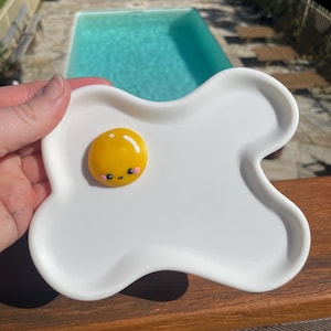May include: A white, egg-shaped dish featuring a yellow, cartoon-style fried egg with a smiling face. The dish is held up against a background of a swimming pool and wooden decking.