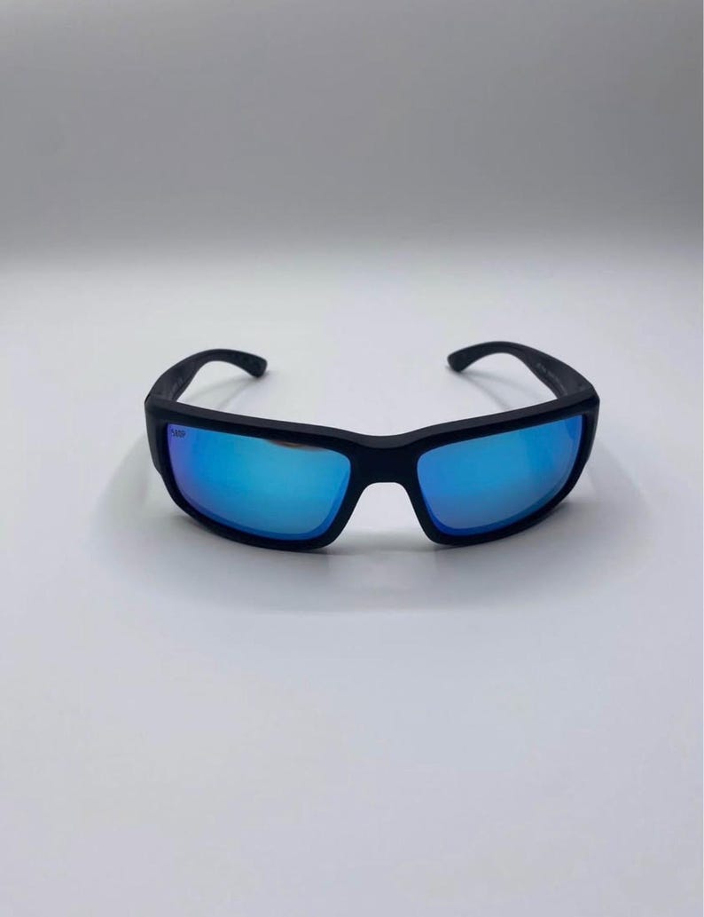 May include: Black sunglasses with blue mirrored lenses. The sunglasses have a rectangular frame and curved temples. The lenses reflect a bright blue color, and the frame appears to be made of a matte black material.