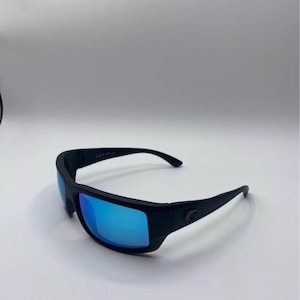 May include: Black sunglasses with blue reflective lenses. The sunglasses have a thick, rectangular frame and curved temples. The lenses reflect a bright blue color. The brand logo is visible on the temple.