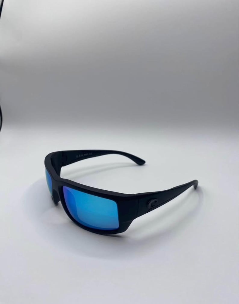 May include: Black sunglasses with blue reflective lenses. The sunglasses have a thick, rectangular frame and curved temples. The lenses reflect a bright blue color. The brand logo is visible on the temple.