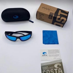 May include: Black sunglasses with blue lenses, a black case, a blue cleaning cloth, and a cardboard box with the text "BORN ON THE WATER" and "COSTA". Also included are two cards with the "COSTA" logo and text.