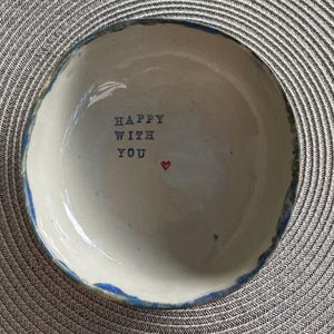 Könnte beinhalten: Eine runde, cremeweiße Keramikschale mit unregelmäßigem, handgeformtem Rand und blauen Akzenten. Auf dem Boden im Inneren befindet sich der schwarze Stempeltext "HAPPY WITH YOU", ergänzt durch ein kleines, handgemaltes rotes Herz.