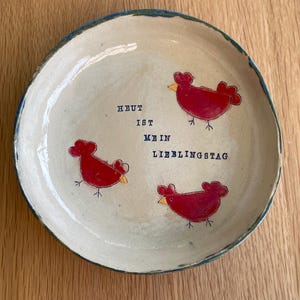 Könnte beinhalten: Handgefertigte Keramikplatte mit cremefarbener Oberfläche und blauem Rand. Die Platte zeigt drei rote Hühnerillustrationen und den deutschen Text "HEUT IST MEIN LIEBLINGSTAG" in Blau.