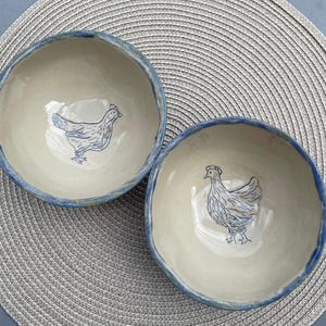 Könnte beinhalten: Zwei runde Keramikschalen mit cremeweißem Innenraum und blau getöntem Rand. Jede Schale zeigt in der Mitte eine handgezeichnete Illustration eines Huhns in blauer Tinte. Die Schalen haben eine organische, leicht unebene Form und Oberfläche.