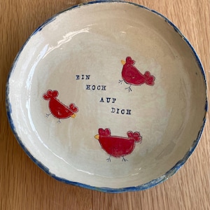Könnte beinhalten: Eine cremefarbene Keramikplatte mit blauem Rand. Sie zeigt drei rote Vogelillustrationen und den deutschen Text "EIN HOCH AUF DICH" in Blau.