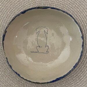 Op de afbeelding: Een handgemaakte keramische kom met een lichtbeige interieur en een donkerblauwe rand. Een eenvoudige omtrek van een hond is blauw getekend in het midden van de kom. De kom is rond en heeft een rustieke, ambachtelijke uitstraling.
