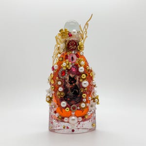 May include: An ornate, decorative bottle with an orange exterior, adorned with pearls, gold accents, and red and clear jewels. The bottle features a small image of two black cats. A clear glass orb sits atop the bottle.