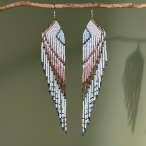 May include: A pair of long, wing-shaped fringe earrings crafted from small glass beads in a palette of white, pale pink, metallic bronze, and light blue. The earrings feature a geometric pattern and hang from gold-toned metal hooks.
