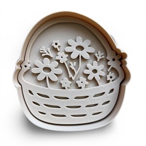 May include: A white plastic cookie cutter shaped like a basket of flowers. The basket has a woven pattern, and the flowers are daisies and small blooms. The cutter is set against a white backdrop.
