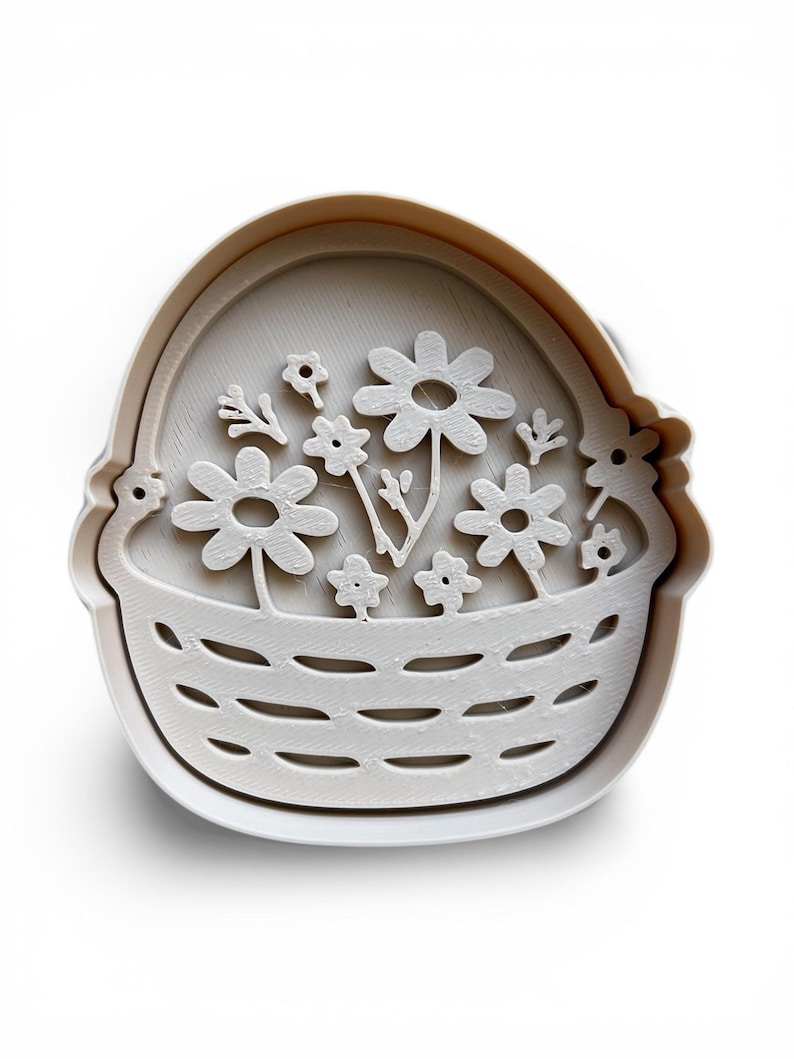 May include: A white plastic cookie cutter shaped like a basket of flowers. The basket has a woven pattern, and the flowers are daisies and small blooms. The cutter is set against a white backdrop.