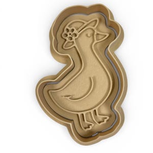May include: A light brown duck-shaped cookie cutter with a floral hat. The cutter has a layered design, with a slightly darker outline. The duck is facing left, with a curved beak and detailed feet. The overall design is simple and elegant.