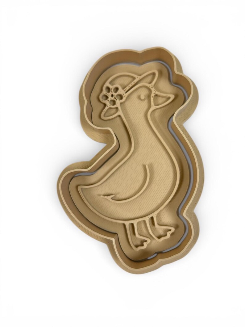 May include: A light brown duck-shaped cookie cutter with a floral hat. The cutter has a layered design, with a slightly darker outline. The duck is facing left, with a curved beak and detailed feet. The overall design is simple and elegant.