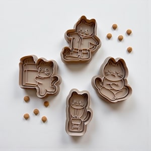 May include: Four tan cat-shaped cookie cutters in different poses. One cat holds a backpack, another stands, and two are sitting. Small, round, brown sprinkles are scattered around the cutters on a white surface.