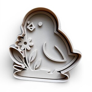 May include: A light grey bird-shaped cookie cutter holding a flower. The cutter has a detailed outline of the bird's body, wings, and a flower with leaves. The design is raised, creating a 3D effect.