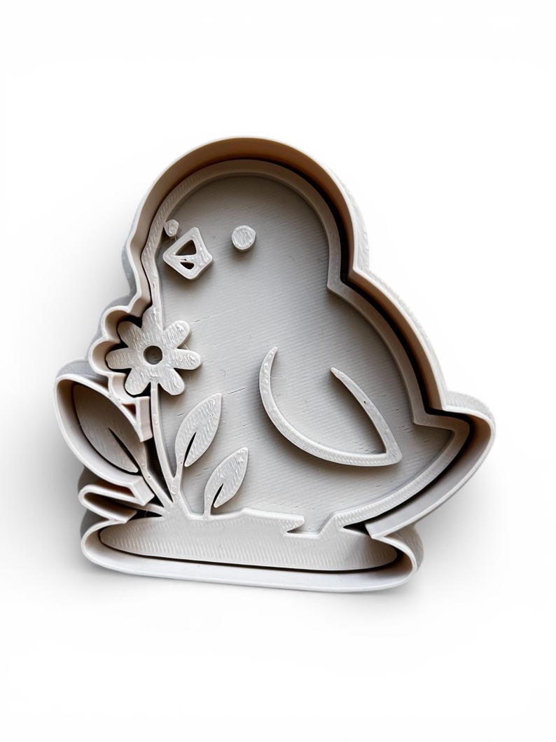 May include: A light grey bird-shaped cookie cutter holding a flower. The cutter has a detailed outline of the bird's body, wings, and a flower with leaves. The design is raised, creating a 3D effect.