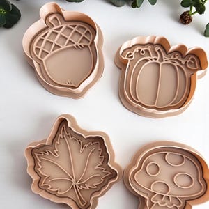 May include: Four tan cookie cutters in autumn shapes: an acorn, a pumpkin, a maple leaf, and a mushroom. The cutters are made of plastic and are arranged on a white surface, with green foliage in the background.