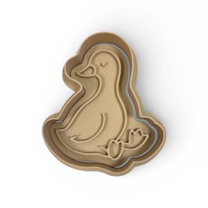 May include: A wooden duck-shaped cookie cutter. The cutter is light brown and features a detailed outline of a duck with a curved neck, eye, and webbed feet. The design is nested within a layered frame.
