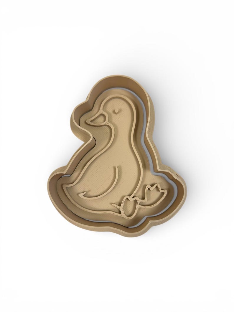 May include: A wooden duck-shaped cookie cutter. The cutter is light brown and features a detailed outline of a duck with a curved neck, eye, and webbed feet. The design is nested within a layered frame.