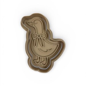 May include: A tan-colored cookie cutter in the shape of a goose with a bow tie. The goose is standing upright with its head tilted slightly. The cutter has a layered design, with a slightly darker outline.