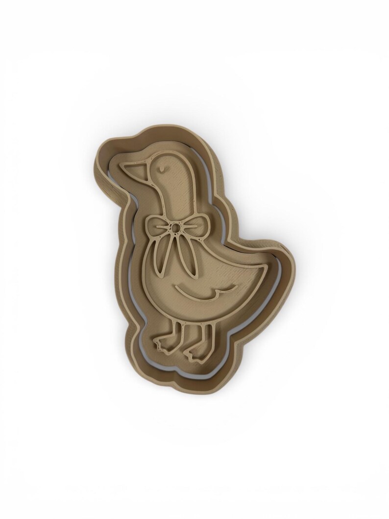 May include: A tan-colored cookie cutter in the shape of a goose with a bow tie. The goose is standing upright with its head tilted slightly. The cutter has a layered design, with a slightly darker outline.