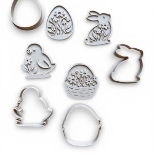 May include: A selection of white Easter cookie cutters. The set features shapes of an egg, a bunny, a chick, a basket of flowers, and other egg and bunny designs. The cutters are made of a light-coloured material and are arranged on a white background.