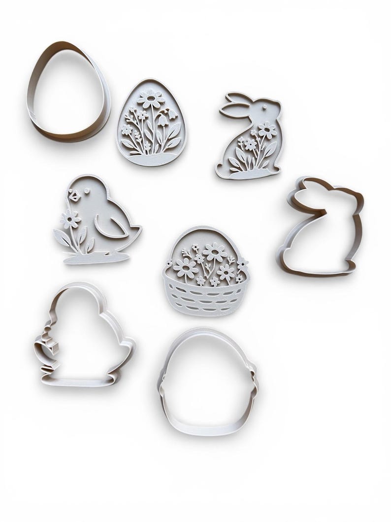 May include: A selection of white Easter cookie cutters. The set features shapes of an egg, a bunny, a chick, a basket of flowers, and other egg and bunny designs. The cutters are made of a light-coloured material and are arranged on a white background.
