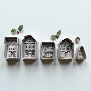 May include: A set of five light grey plastic cookie cutters featuring four distinct house designs and one tree shape, accompanied by two small sprigs of green leaves with red berries, arranged in a horizontal line on a plain white surface.