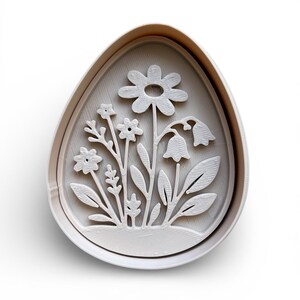 May include: A white, egg-shaped cookie cutter with a floral design. The design includes a large daisy, bell-shaped flowers, and various leaves and stems. The cutter is made of plastic and is for baking.