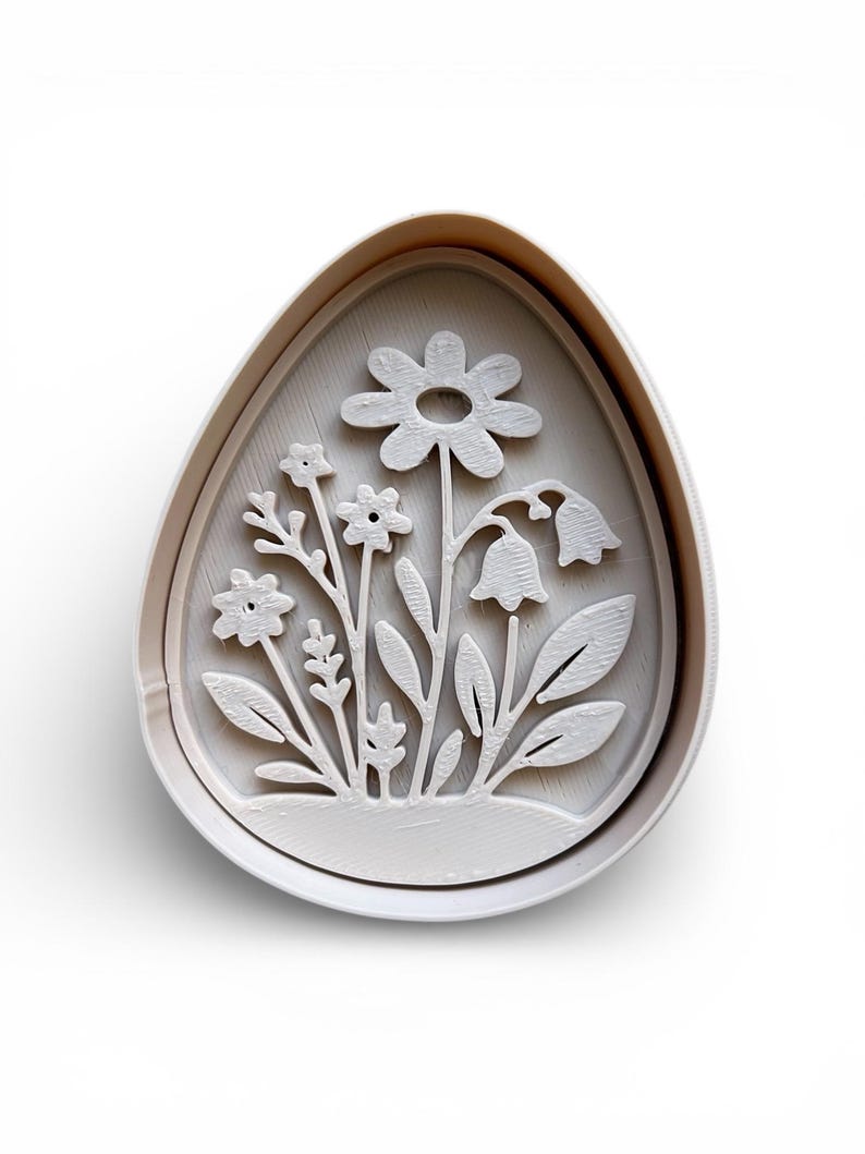 May include: A white, egg-shaped cookie cutter with a floral design. The design includes a large daisy, bell-shaped flowers, and various leaves and stems. The cutter is made of plastic and is for baking.