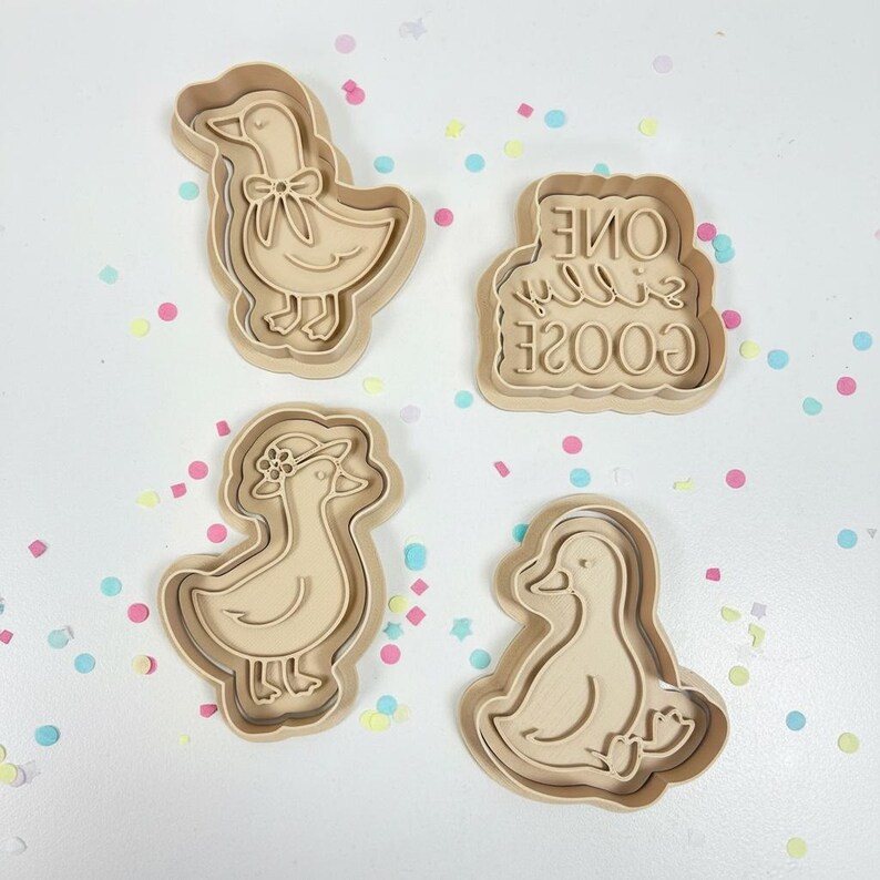 May include: Four beige cookie cutters in the shape of geese. One goose has a bow, another has a hat, and a third has the text "ONE SILLY GOOSE". The cookie cutters are surrounded by colorful confetti.