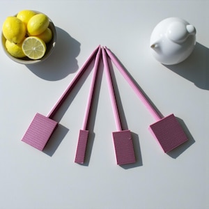 May include: Four pink, rectangular, textured objects with long, thin handles are arranged on a white surface. A bowl of lemons and a white bird-shaped ornament are also present. The objects appear to be decorative or functional.