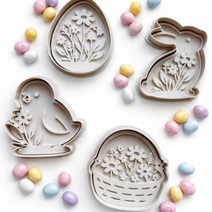 May include: A collection of Easter cookie cutters shaped like an egg, bunny, chick, and basket, all with floral patterns. Pastel-coloured speckled eggs are scattered around the cutters on a white background.