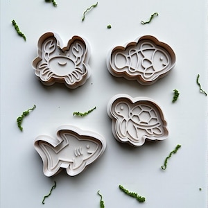May include: Four white plastic cookie cutters shaped like sea creatures: a crab, a fish, a shark, and a turtle. Green decorative sprigs are scattered around the cutters on a white surface. The cutters are designed with detailed outlines.