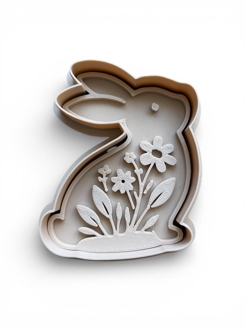 May include: A white, bunny-shaped cookie cutter with a floral design. The cutter features a detailed outline of a rabbit with flowers and leaves inside. The design is raised, creating a 3D effect. Ideal for Easter or spring-themed baking.