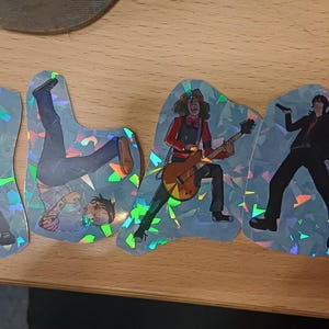 Puede incluir: Cuatro pegatinas holográficas con ilustraciones de músicos en varias poses. Una figura sostiene una guitarra, otra un micrófono y una tercera está boca abajo. Las pegatinas tienen un efecto brillante e iridiscente.