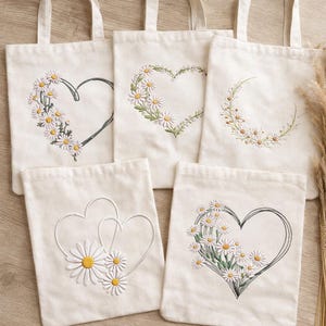 Op de afbeelding: Vijf ecru canvas draagtassen met madeliefjes hart ontwerpen. Elke tas heeft een ander geborduurd hartvorm, sommige omlijnd en andere gevuld met witte madeliefjes en gele centra. De tassen hebben bijpassende ecru handvatten.