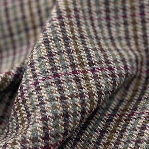 Puede incluir: Primer plano de una tela de tweed doblada. El material presenta un patrón tradicional en tonos verde oliva, marrón, crema y burdeos. La textura tejida es evidente, con un tejido diagonal que crea una apariencia texturizada. La tela está doblada.