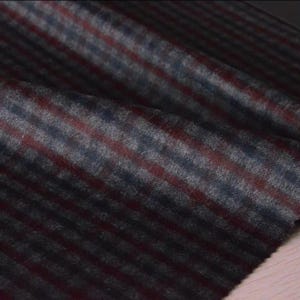 May include: Close-up of a folded fabric with a plaid pattern. The fabric features a combination of dark grey, burgundy, and blue stripes. The material appears to be soft and textured, with a subtle sheen.