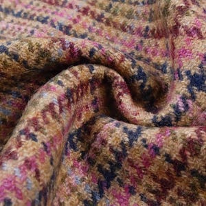 May include: Close-up of a textured fabric with a houndstooth pattern. The fabric features a blend of colors, including tan, brown, pink, blue, and green. The material is folded, creating shadows and depth, highlighting the woven texture.