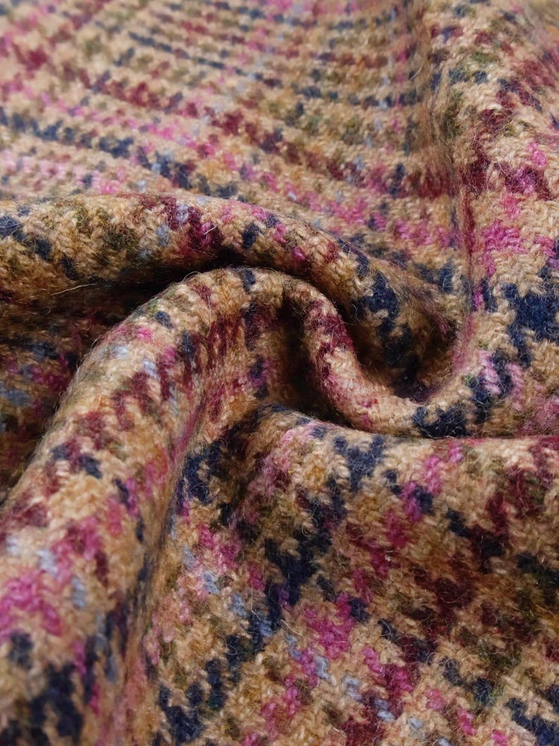 May include: Close-up of a textured fabric with a houndstooth pattern. The fabric features a blend of colors, including tan, brown, pink, blue, and green. The material is folded, creating shadows and depth, highlighting the woven texture.