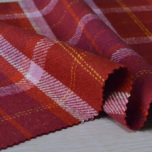 May include: Close-up of a folded piece of red plaid fabric. The fabric has a pattern of red, white, and yellow lines. The fabric is soft and appears to be wool or a similar material. The edges of the fabric are visible.