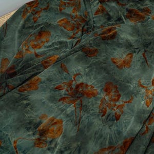 Op de afbeelding: Close-up van een teal stof met een bloemen- en vlinderpatroon in roest- en bruintinten. De stof heeft een zachte, fluweelachtige textuur met een subtiel tie-dye effect, wat diepte en visuele interesse creëert.