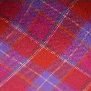 May include: Close-up of a vibrant plaid fabric in shades of red, pink, and purple. The pattern features intersecting lines of white, orange, and teal, creating a classic tartan design. The fabric has a soft, textured appearance.