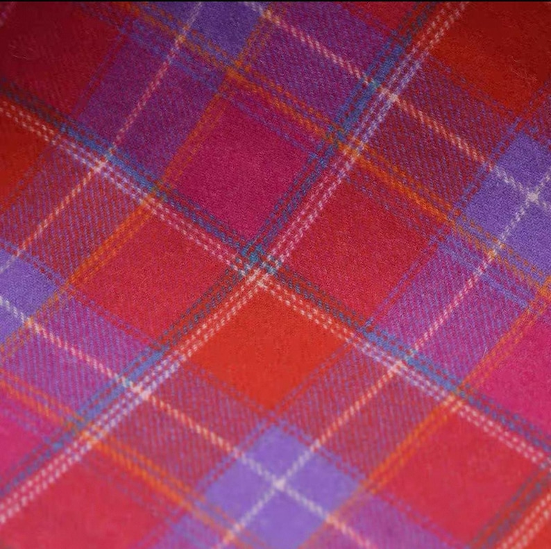 May include: Close-up of a vibrant plaid fabric in shades of red, pink, and purple. The pattern features intersecting lines of white, orange, and teal, creating a classic tartan design. The fabric has a soft, textured appearance.