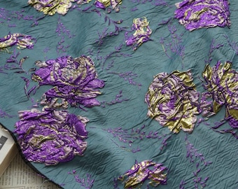 May include: Close-up of teal fabric with raised floral embroidery in purple and gold. The textured material features large, detailed flowers and delicate vines, creating a luxurious and ornate design. Ideal for apparel or home decor.