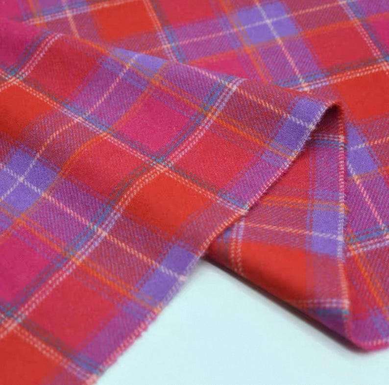 May include: A close-up shot of a colourful plaid fabric, predominantly red, pink, and purple. The tartan pattern is formed by intersecting lines, creating a grid. The fabric is folded, revealing its texture and the interplay of colours.