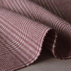 May include: Close-up of a folded fabric with a plaid pattern in shades of burgundy and white. The fabric has a textured appearance and a visible selvedge edge. The plaid design features intersecting lines creating a grid-like pattern, suitable for apparel or home decor.