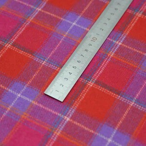 May include: A close-up shot of a colourful plaid fabric in red, pink, and purple hues. A metal ruler, marked in centimetres, is placed diagonally across the fabric. The fabric's texture is visible, suggesting a woven material.