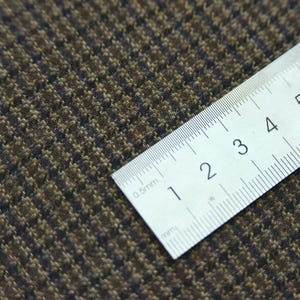 May include: Close-up of brown and black patterned fabric with a ruler showing measurements in millimeters. The fabric has a textured appearance with a houndstooth design. The ruler is white with black markings.