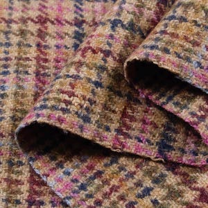 May include: Close-up of a folded fabric with a houndstooth pattern. The fabric features a blend of colors, including tan, pink, blue, and burgundy. The texture appears to be a woven tweed, suitable for apparel or home decor.